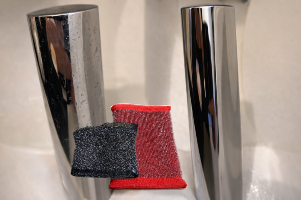 洗面台のクローム水栓の横に黒と赤の金属メッシュスポンジを置いた様子。金属部分の反射とスポンジの質感が分かる写真。掃除のビフォーアフター
