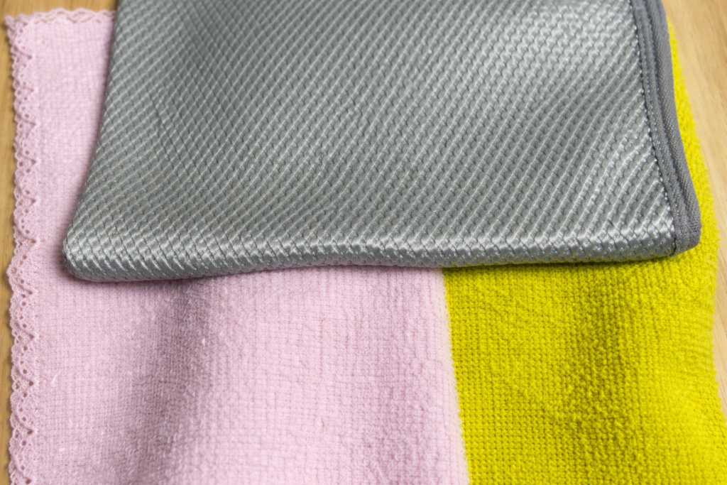 ピンクと黄色のマイクロファイバークロスの上に、グレーの凹凸加工クロスを重ねて並べた写真。素材や質感の違いが分かる構図。