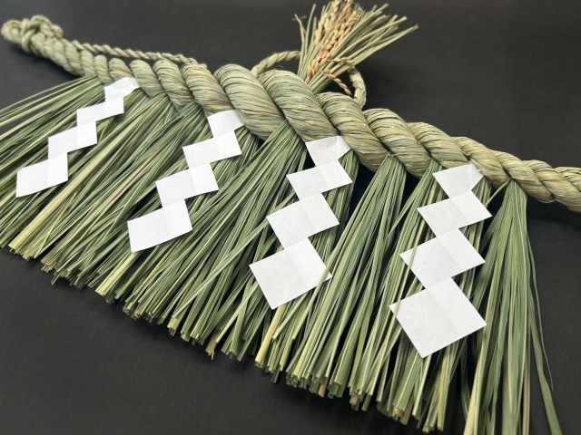 緑色の稲わらで作られたしめ縄飾り。白い紙垂（しで）が下がった状態で、黒い背景の上に置かれている様子。