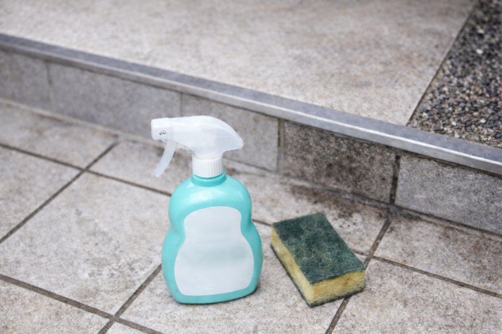 玄関タイルの上に置いたウタマロクリーナーのスプレーボトルと、使い古したキッチンスポンジ。屋外タイルの黒ずみ掃除に使う道具を並べた写真。